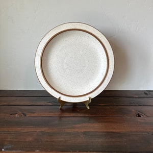May include: A white ceramic dinner plate with a brown rim and speckled glaze. The plate is on a wooden stand.