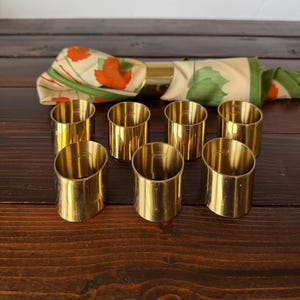 May include: Eight gold-colored napkin rings are arranged on a dark wooden surface. A folded napkin with orange and green leaf patterns is visible in the background. The napkin rings are cylindrical and have a polished finish.