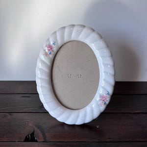 May include: White oval ceramic picture frame with a rope-like design around the edge. The frame has a floral design with pink and blue flowers.