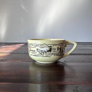 May include: A light yellow ceramic mug with a black and white landscape design. The mug has a rounded shape with a handle on the right side. The landscape design features trees, horses, and a person riding a horse.