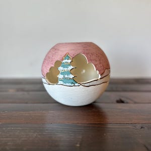 May include: A spherical ceramic vase with a textured pink upper half and a white lower half. The design features a stylized landscape with a teal tree and gold-colored clouds. The vase sits on a dark wooden surface.