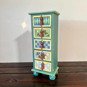 May include: A small, teal-colored wooden cabinet with five drawers. Each drawer is decorated with a different pattern and a small, red umbrella-shaped handle. The cabinet sits on four blue, polka-dotted feet.