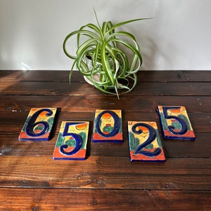 May include: Set of five ceramic house numbers with a southwestern design. The numbers are 6, 5, 0, 2, and 5. Each number is painted with a yellow sun, blue sky, green cactus, and brown desert landscape.