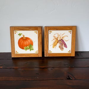 May include: Two square wall plaques featuring ceramic tiles set in light oak wood frames. One tile displays a vibrant orange pumpkin with green leaves, while the other tile depicts a cluster of multi-colored Indian corn with dried husks.