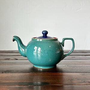 May include: A teal ceramic teapot with a rounded body, a curved spout, and a handle. The lid is dark blue with a small knob. The teapot sits on a dark wooden surface. The teapot has a speckled texture.