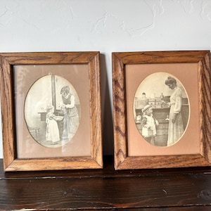 May include: Two framed vintage prints with oval-shaped images. The prints are in wooden frames with a light brown matting. The images are black and white, depicting scenes of a woman and child. One shows a woman and child near a stove, the other shows a woman and child near a counter.