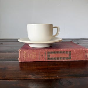 May include: A cream-colored teacup and saucer set on top of a vintage book with a red cover. The teacup has a simple, rounded design with a handle. The saucer is circular, and the set is placed on a book with the title "THE YELLOW HOUSE".