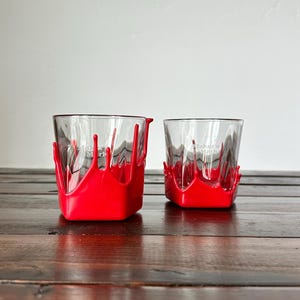 May include: Two clear square-shaped glasses with a red paint drip design. The glasses have the text "Maker's Mark" printed on them. The glasses are sitting on a dark wooden surface.