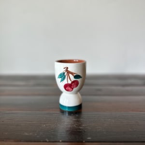 May include: White ceramic egg cup with a brown rim and a green and red cherry design.