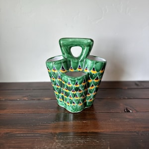 May include: A green ceramic utensil holder with a handle. The holder has three compartments and is decorated with a repeating pattern of yellow, blue, and green scales. The holder is sitting on a dark wooden surface.