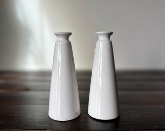 May include: Two white ceramic vases with a conical shape. The vases are sitting on a dark wooden surface.