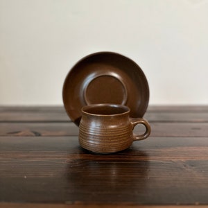May include: A brown ceramic coffee cup and saucer set. The cup has a textured, ribbed design and a rounded handle. The saucer is plain and has a slightly raised lip.