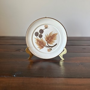 May include: A white ceramic plate with a brown floral design. The plate has a gold rim and is on a gold metal stand.