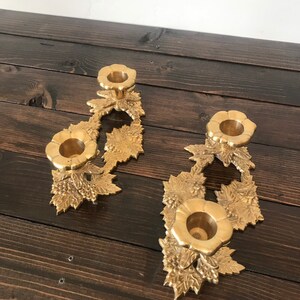 May include: A pair of gold metal candle holders with a floral design. Each holder has three candle cups and features a grapevine motif.