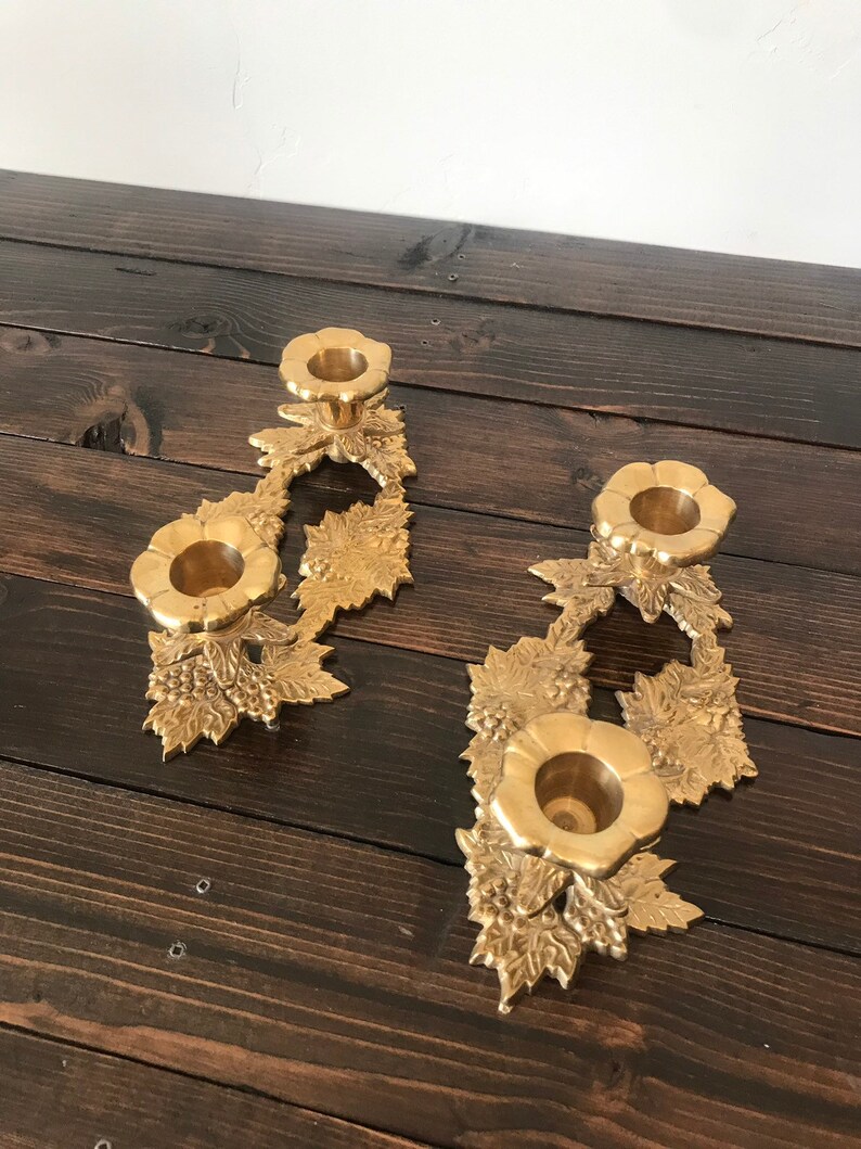 May include: A pair of gold metal candle holders with a floral design. Each holder has three candle cups and features a grapevine motif.