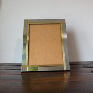 May include: A rectangular silver-toned picture frame with gold-colored trim. The frame has a simple, elegant design with a neutral-colored backing. The frame is displayed on a dark wooden surface.