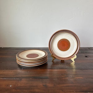 May include: A stack of five brown and white ceramic plates with a red circle design in the center. The plates are on a dark brown wooden surface. One plate is on a gold metal stand.