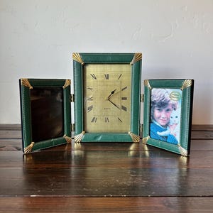 May include: A set of three vintage green leatherette picture frames and a clock. The clock has a gold face with Roman numerals. The frames have gold corner accents and are hinged together. One frame holds a photo of a child.