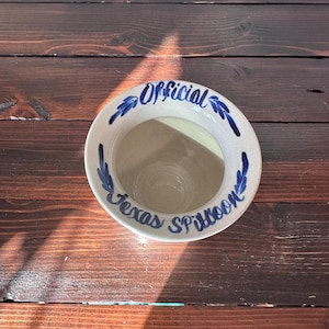 May include: A white ceramic bowl with blue and white floral designs. The bowl has the words "Official Texas Spittoon" written on it.