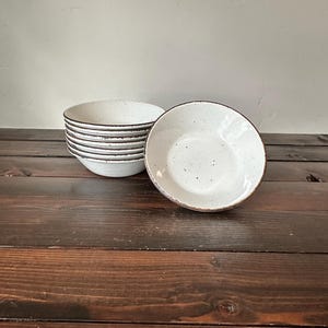 May include: A stack of white ceramic bowls with a speckled design and a dark brown rim. One bowl is separate, showing the interior. The bowls are on a dark brown wooden surface.