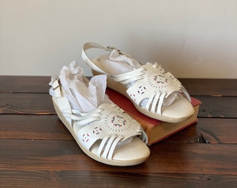 May include: A pair of white leather sandals with a low wedge heel. The sandals feature a strappy design with decorative cutouts and a buckle closure. The shoes are resting on a vintage book on a dark wooden surface.