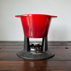 May include: A bright red enameled cast iron fondue pot with two side handles, resting on a black metal stand. The base features a circular burner component with a small silver-toned metal handle, designed to hold a heat source beneath the pot.