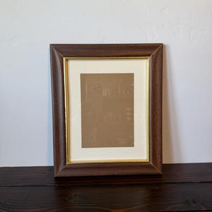 May include: A rectangular picture frame with a dark brown wood-grain finish and a thin gold inner border. The frame includes an off-white mat board surrounding a central rectangular opening that displays a plain brown backing material.
