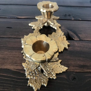 May include: A gold metal candle holder with a floral design. The holder has two candle cups and is decorated with leaves and grapes.