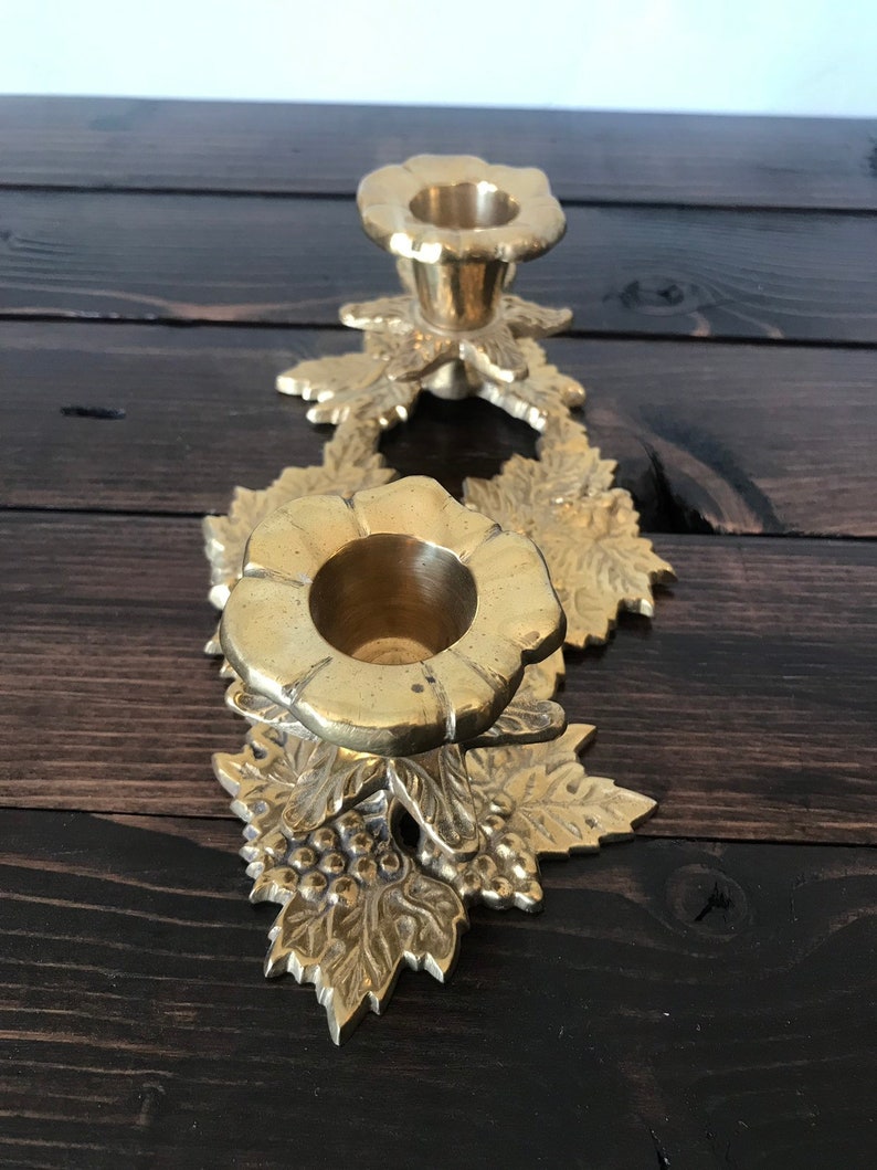 May include: A gold metal candle holder with a floral design. The holder has two candle cups and is decorated with leaves and grapes.
