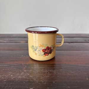 May include: A vintage yellow enamel mug with a floral design featuring red and white roses. The mug has a brown rim and handle. The background is a wooden surface.