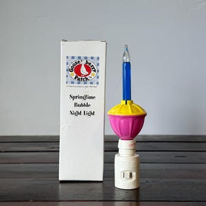 May include: A Gooseberry Patch Springtime Bubble Night Light with a white box featuring a blue plaid logo and text. The night light has a white plug base, a pink and yellow plastic housing, and a tall, thin, blue glass bubble light bulb on top.