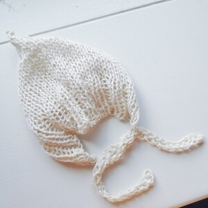 Könnte beinhalten: Ein weißes gehäkeltes Baby-Mützchen mit einer langen Schleife. Die Mütze ist aus weichem, leichtem Garn gefertigt und hat eine zarte, spitzenartige Textur.