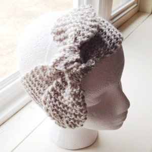 May include: A beige knitted headband with a folded design. The headband is displayed on a white mannequin head.