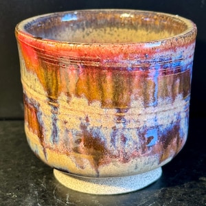 Puede incluir: Una taza de cerámica con un esmalte marrón, rojo y azul. La taza tiene una base ancha y un cuerpo redondeado.