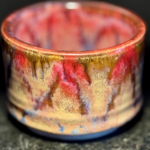 Puede incluir: Un pequeño cuenco de cerámica con un esmalte rojo, amarillo y marrón. El esmalte tiene un aspecto moteado, con vetas de color que lo atraviesan.