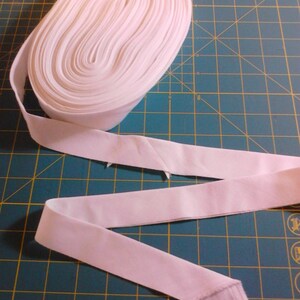 May include: A white cotton ribbon rolled up on a green cutting mat. The ribbon is approximately 1 inch wide and is shown unfurled in a loose S shape.