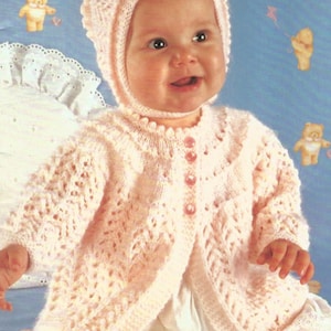 May include: A pale pink knitted baby cardigan featuring a delicate openwork lace pattern, long sleeves, and a round neckline with three translucent pink buttons down the front, paired with a matching knitted bonnet with a scalloped edge.