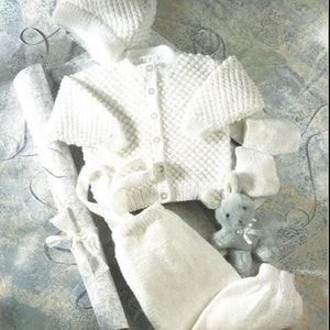 May include: A white knitted baby set consisting of a long-sleeved button-down cardigan with a textured popcorn stitch pattern, matching trousers, a beanie hat, and a pair of mittens. A small, light blue plush rabbit toy is positioned nearby.