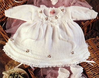 May include: A white knitted baby dress with long sleeves, a ruffled collar, and small pink floral ribbon accents. The set includes a matching white knitted bonnet and a pair of white knitted booties, all featuring delicate ribbon ties and lace trim.