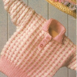 Puede incluir: Un suéter tejido de manga larga con un patrón de textura de gofre en hilo rosa y blanco alternado, un cuello con tres botones pequeños y un dobladillo acanalado. Un sombrero de paja decorado con flores rosas y blancas se sitúa encima.