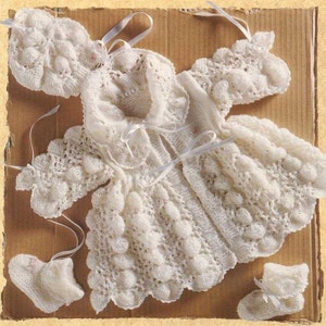May include: A white knitted baby set consisting of a long-sleeved cardigan with a textured bobble pattern, a matching bonnet, and a pair of booties. The cardigan features a ruffled collar and thin white ribbon ties at the front closure.