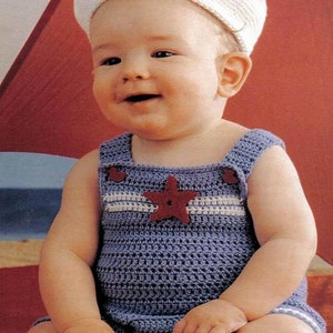 May include: A white crocheted cap and a blue crocheted romper featuring a horizontal white stripe across the chest and a dark red star appliqué centred on the front. The romper has wide shoulder straps and is constructed with a textured stitch.