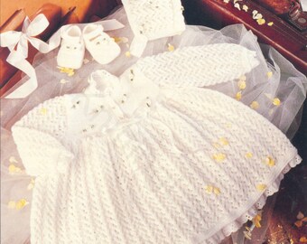 May include: A white knitted baby dress with long sleeves, a matching bonnet, and a pair of booties. The dress features a textured chevron pattern, a ribbon tie at the waist, and a delicate lace trim along the hem and neckline, all in white.