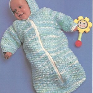 May include: A hooded baby bunting bag made of variegated light blue, white, and green yarn with a central white zipper closure. The garment features long sleeves and a textured knit pattern. A yellow plastic flower-shaped rattle is positioned nearby.