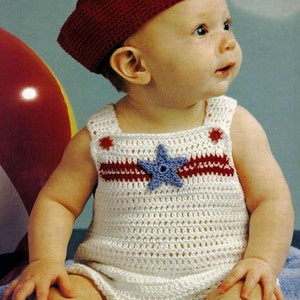 May include: A white crocheted romper featuring a horizontal red stripe across the chest, a blue star appliqué, and small red circular accents at the shoulder straps, paired with a solid dark red crocheted cap with a flat top and structured brim.