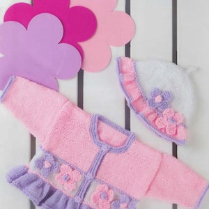 Pode incluir: Um conjunto de malha para bebé composto por um casaco e um gorro a condizer em rosa, branco e lavanda. O casaco apresenta uma bainha com folhos lavanda, acabamentos lavanda e aplicações florais. O gorro branco tem uma aba com folhos lavanda.