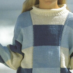 Puede incluir: Un suéter de punto de manga larga con un diseño de bloques grandes en tonos azul claro, azul medio y crema. La prenda presenta un cuello alto acanalado y puños acanalados a juego, confeccionado con un punto de jersey visible.