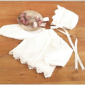 May include: A white knitted cardigan with a scalloped hem and matching bonnet featuring long ribbon ties. A small, round, woven basket decorated with pink flowers and a light pink ribbon bow is positioned near the cardigan and bonnet set.