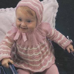 May include: A knitted baby outfit consisting of a pink and white striped bonnet with a pink ribbon tie, a matching long-sleeved sweater with a flared, textured skirt, and solid pink leggings. The sweater features horizontal stripes and ribbon accents.