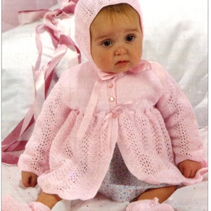 May include: A light pink knitted baby cardigan featuring a delicate openwork pattern, long sleeves, and a front button closure with a matching satin ribbon tie. The set includes a coordinating knitted bonnet and booties, all in a soft pastel pink.